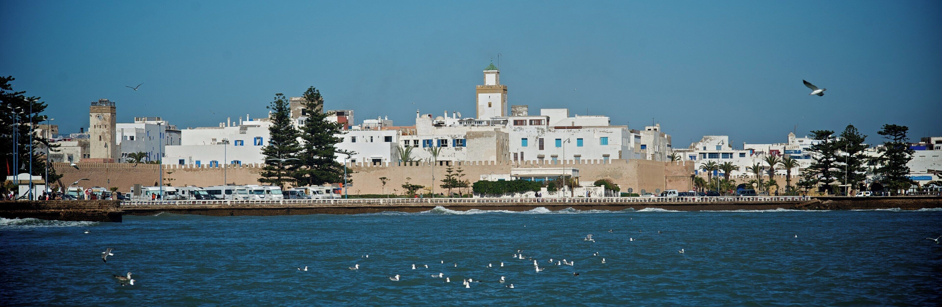 Paisagem costeira com edifícios históricos, uma torre imponente e um calçadão à beira-mar.
