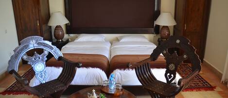 A hotel room with a large bed, two nightstands with lamps, a wooden coffee table with a teapot and a bottle, and two ornate chairs.