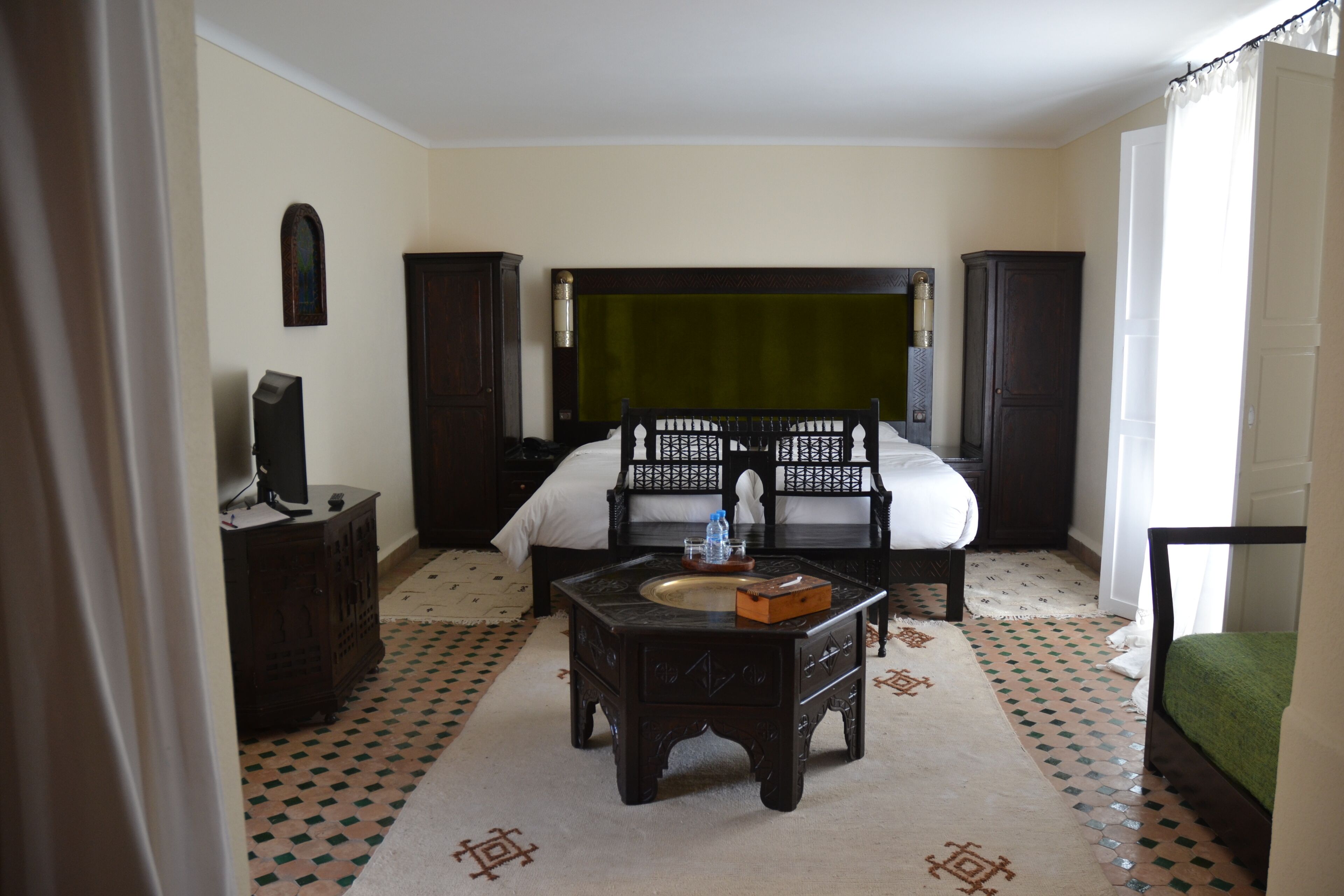 A room with a bed, a wooden cabinet, a small table with a basin, and patterned flooring.
