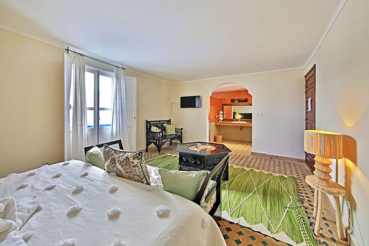 A hotel room with a bed, a small table, a chair, a TV, and a view of the kitchen and dining area.