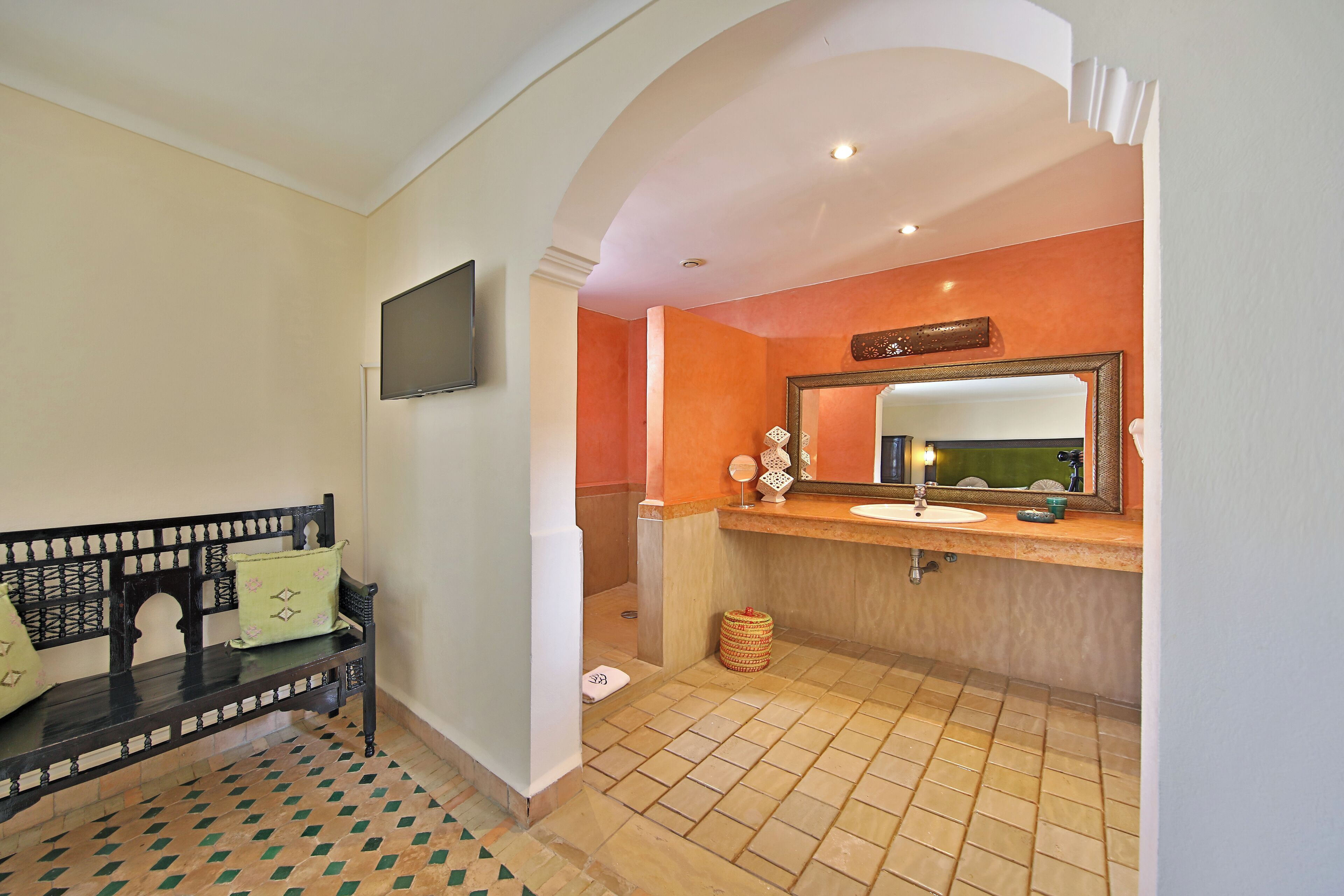 A bathroom with a bench, a sink, a mirror, and a television mounted on the wall.