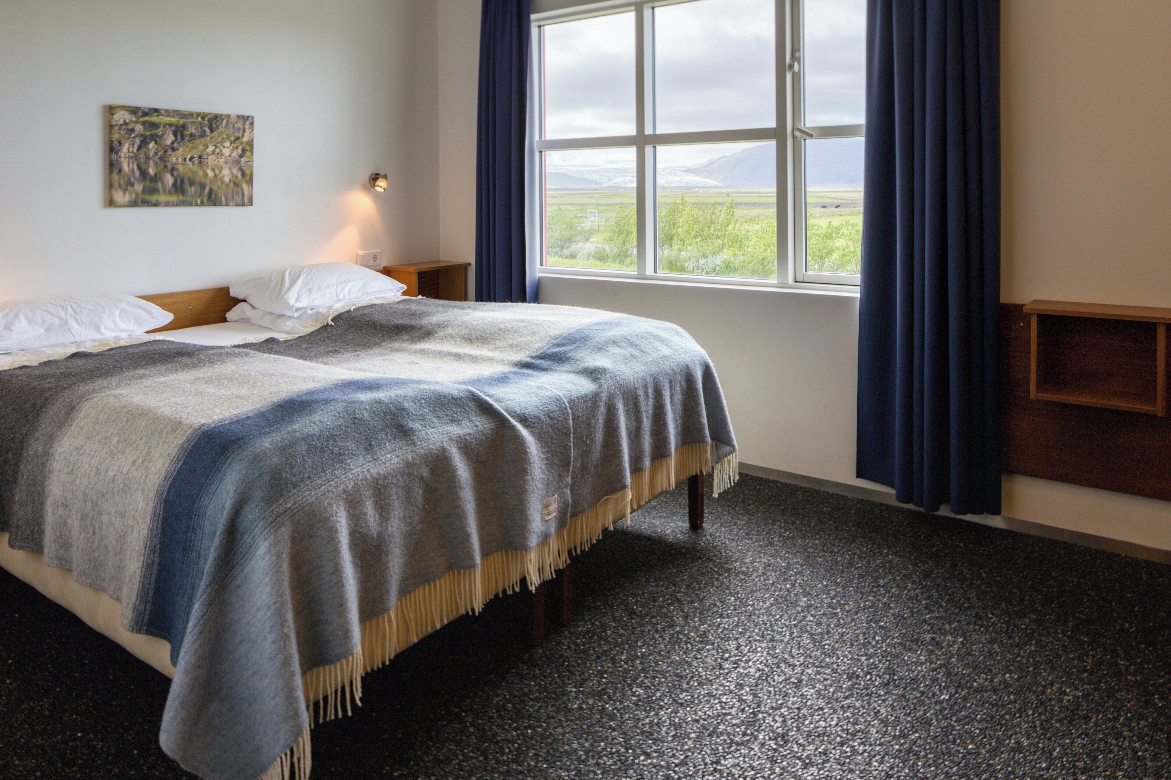 A bedroom with a bed, a nightstand, a window with curtains, a painting on the wall, and a view of the outdoors.