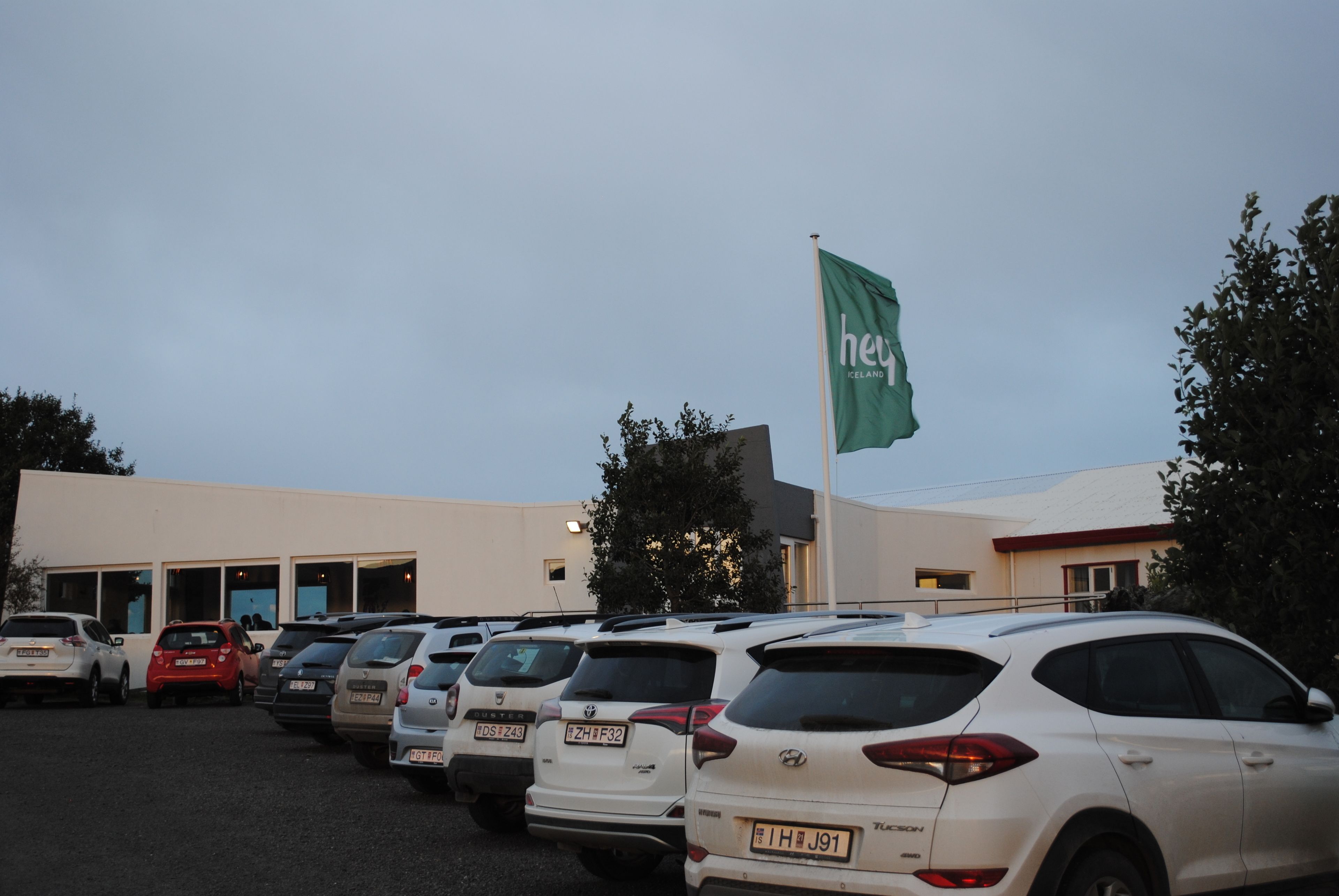 Um estacionamento com vários carros, um edifício com uma bandeira verde e árvores ao fundo.