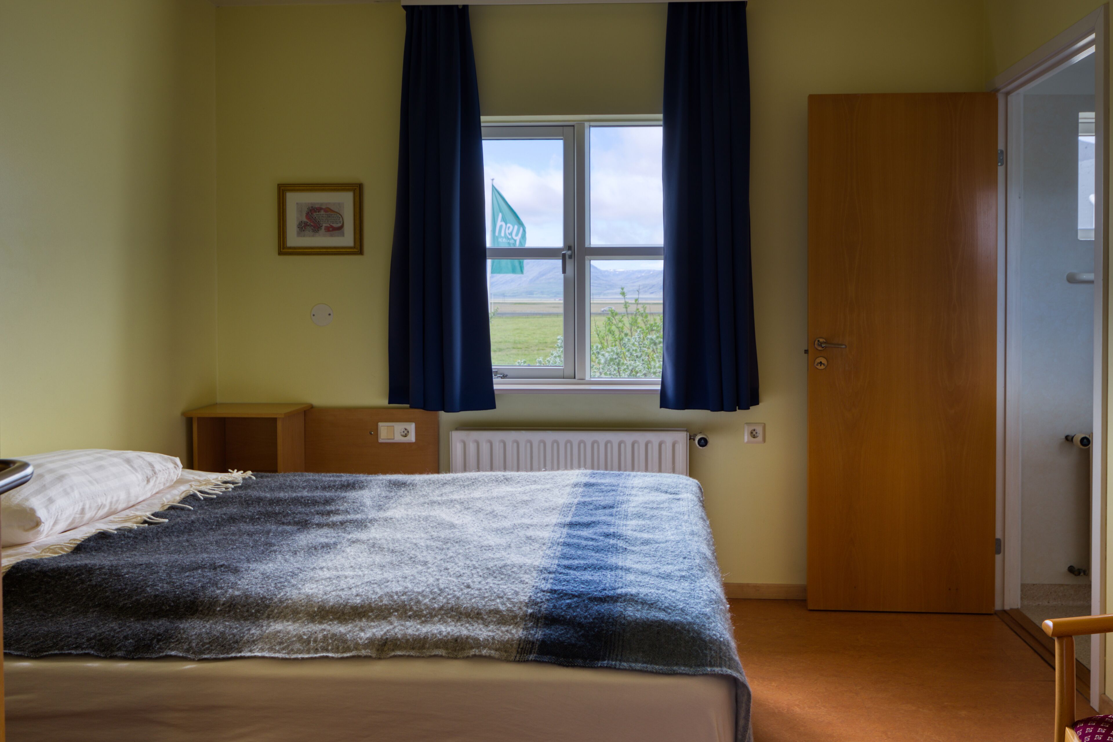 A bedroom with a bed, a window with curtains, a radiator, and a door.