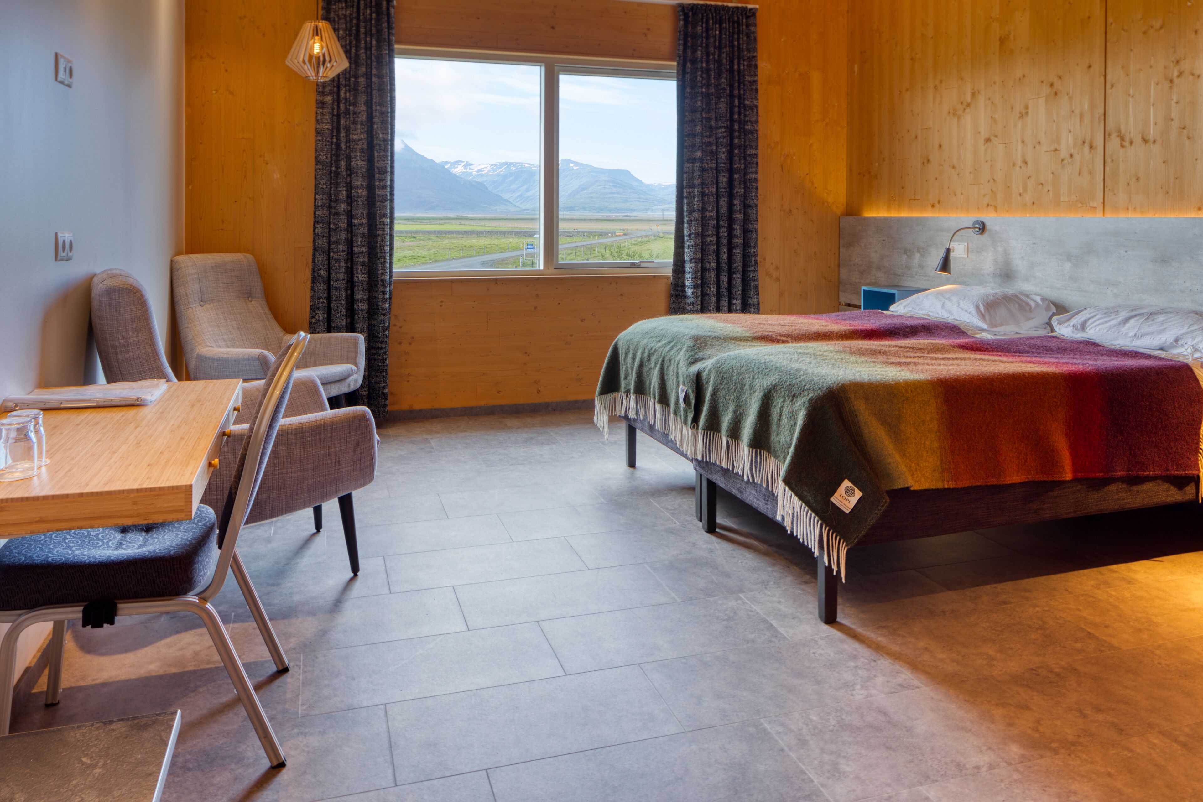 A room with a bed, a desk, and two chairs. There is a window with a view of mountains.