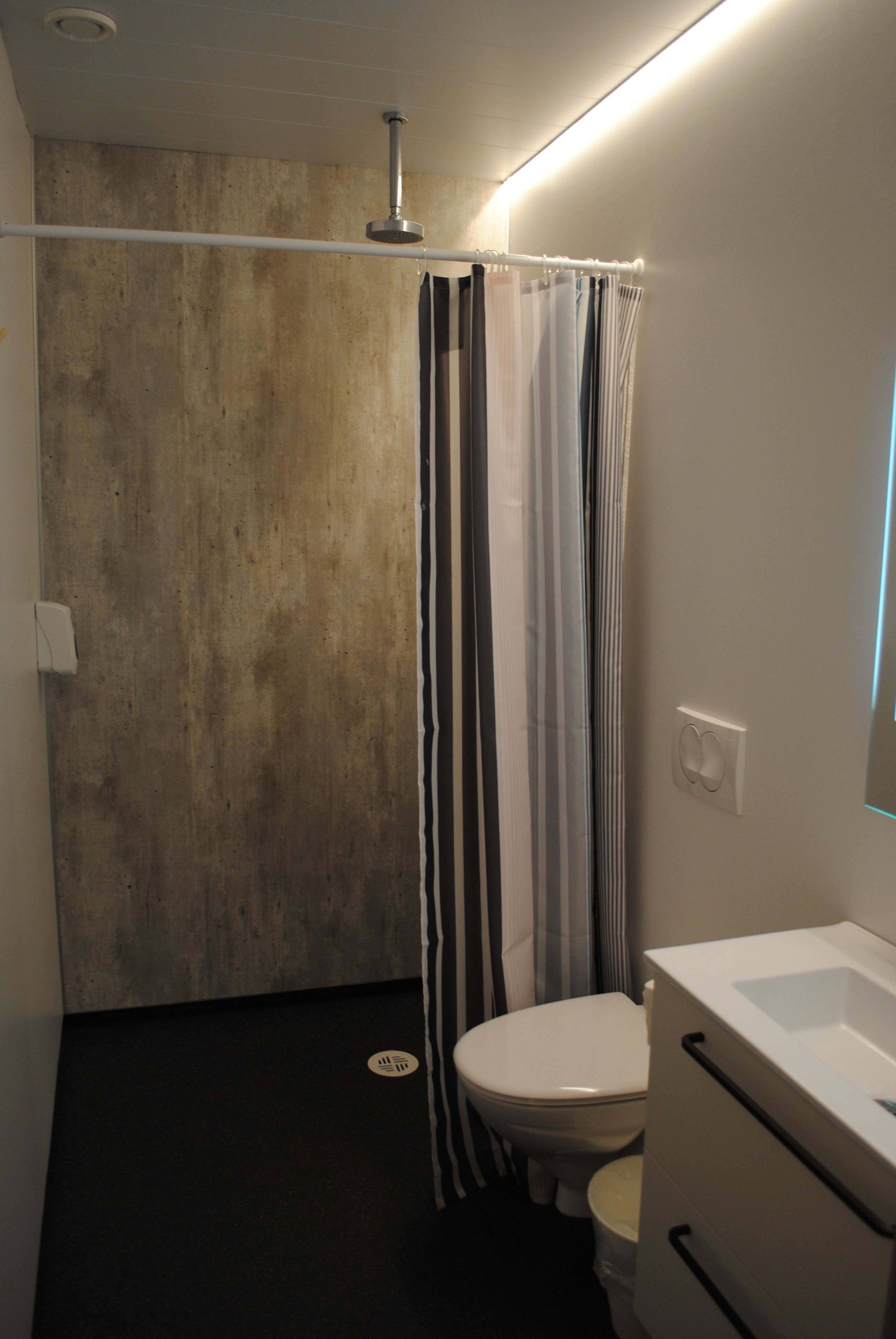 Un bagno con parete in cemento, una doccia con tende a righe, un WC bianco e un lavandino bianco con uno specchio.
