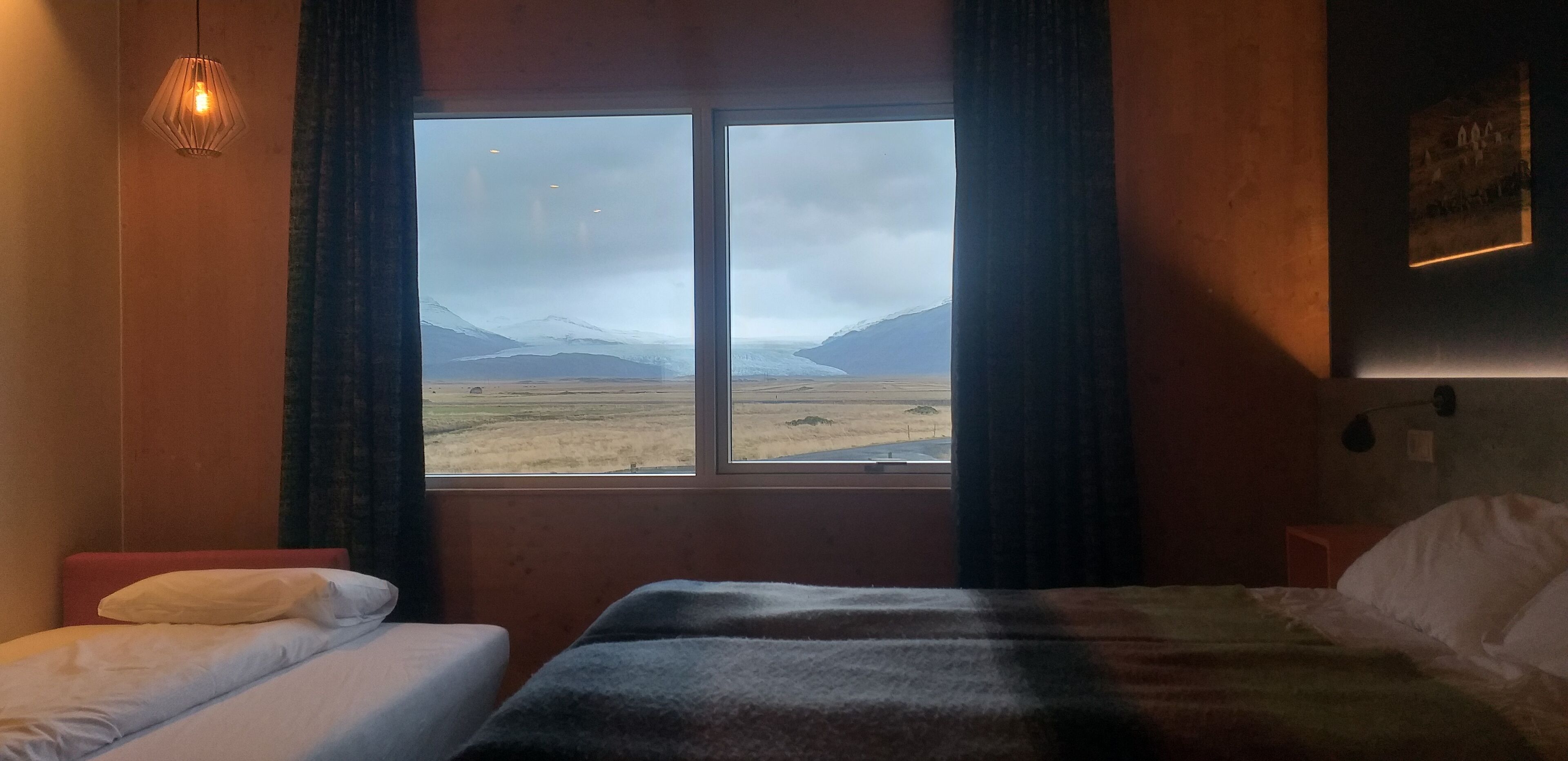A hotel room with two beds, a large window with a view of mountains and a cloudy sky, and a wall-mounted light fixture.