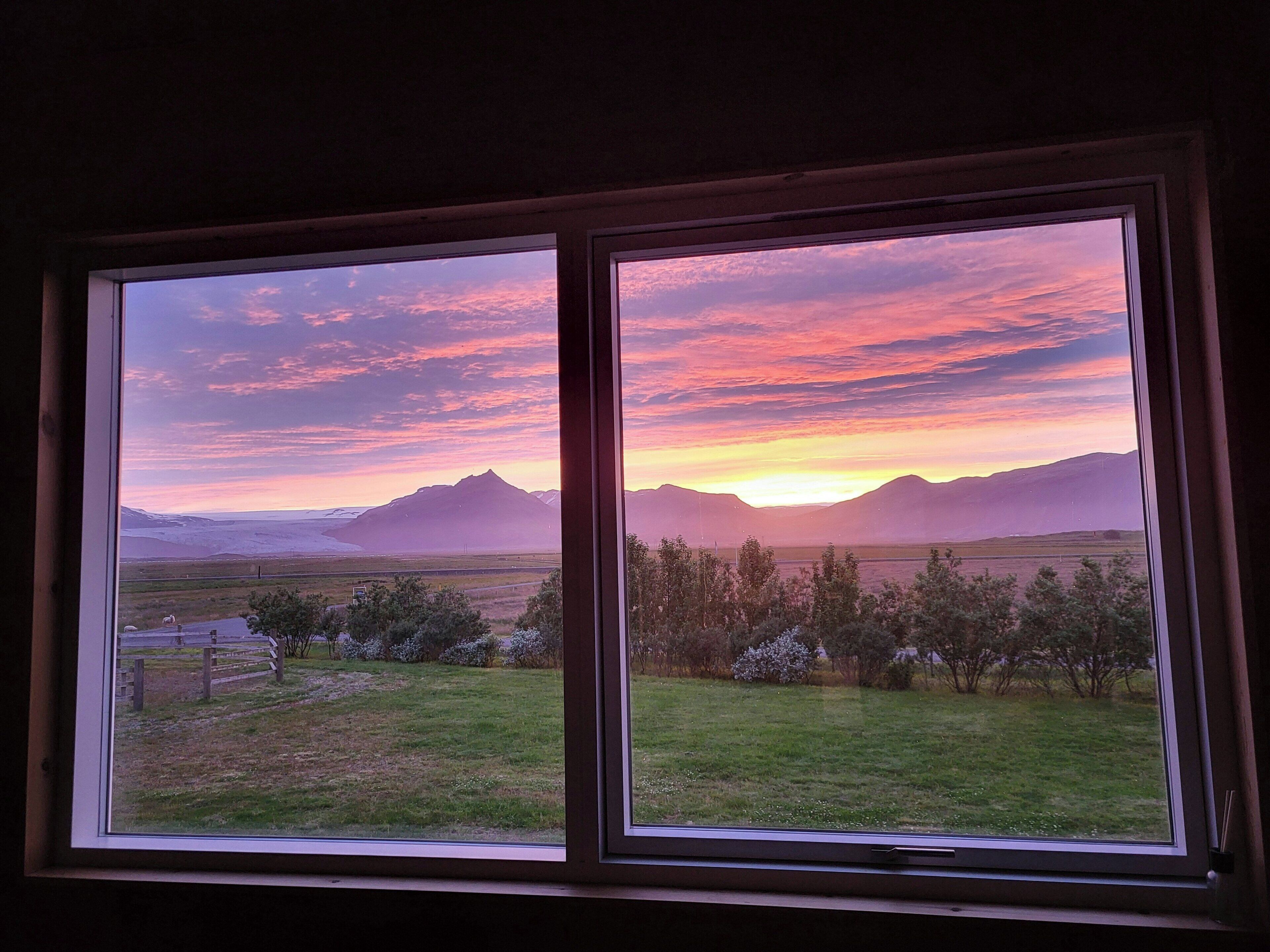 A window with a view of a sunset over mountains and a grassy field.