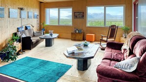 A wooden interior with a sofa, a coffee table, and a chair. There are windows with a view of mountains. - Brunnhóll Country Guesthouse (Höfn)