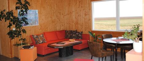 A cozy room with a wooden wall, a red sofa, a coffee table, a dining table with chairs, and a large window offering a view of the outdoors.