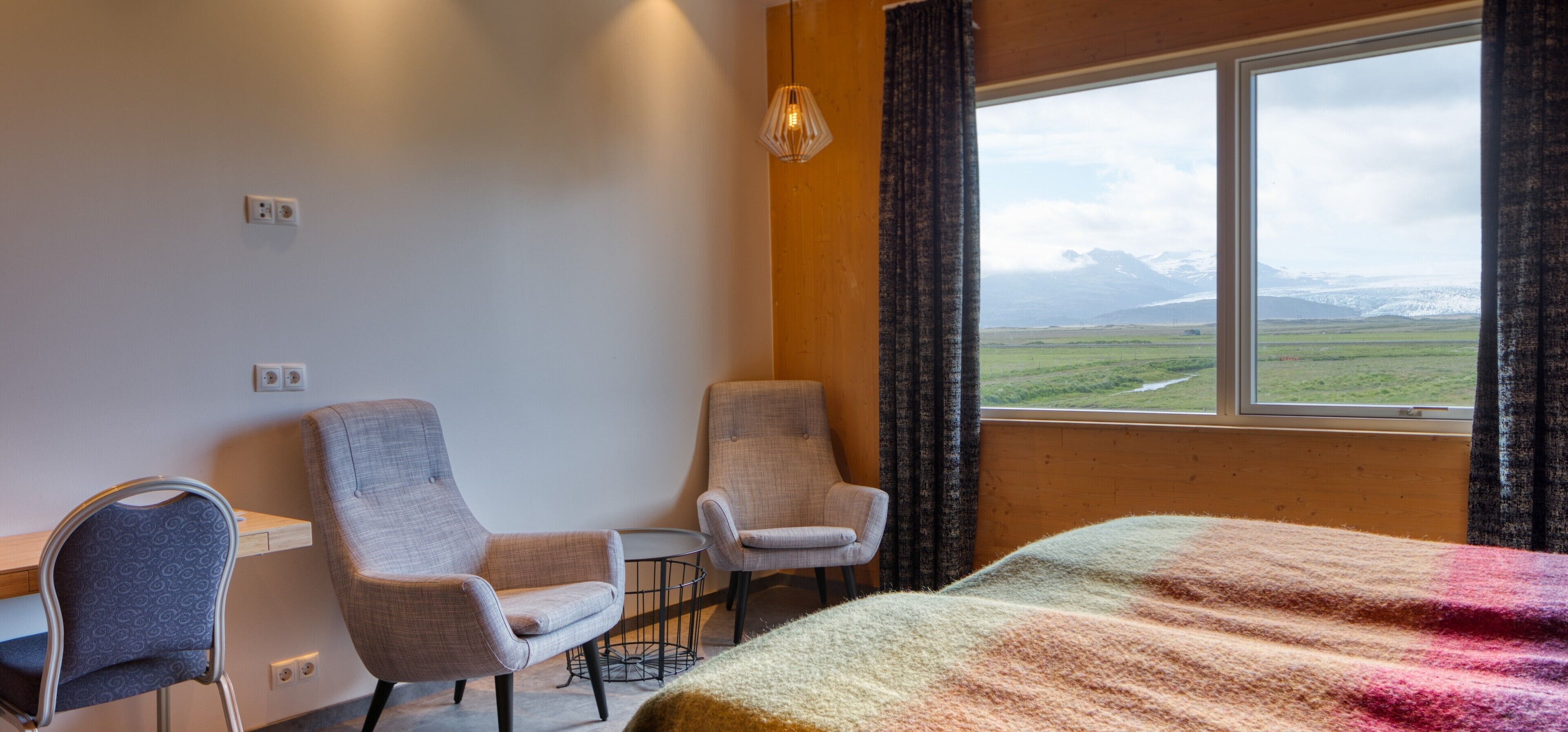 A room with two armchairs, a small table, a bed, and a window with a view of mountains.