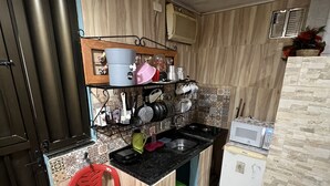 Uma cozinha compacta com bancada preta, pia, micro-ondas e geladeira. Há uma cadeira de plástico vermelha e uma pequena lixeira verde.