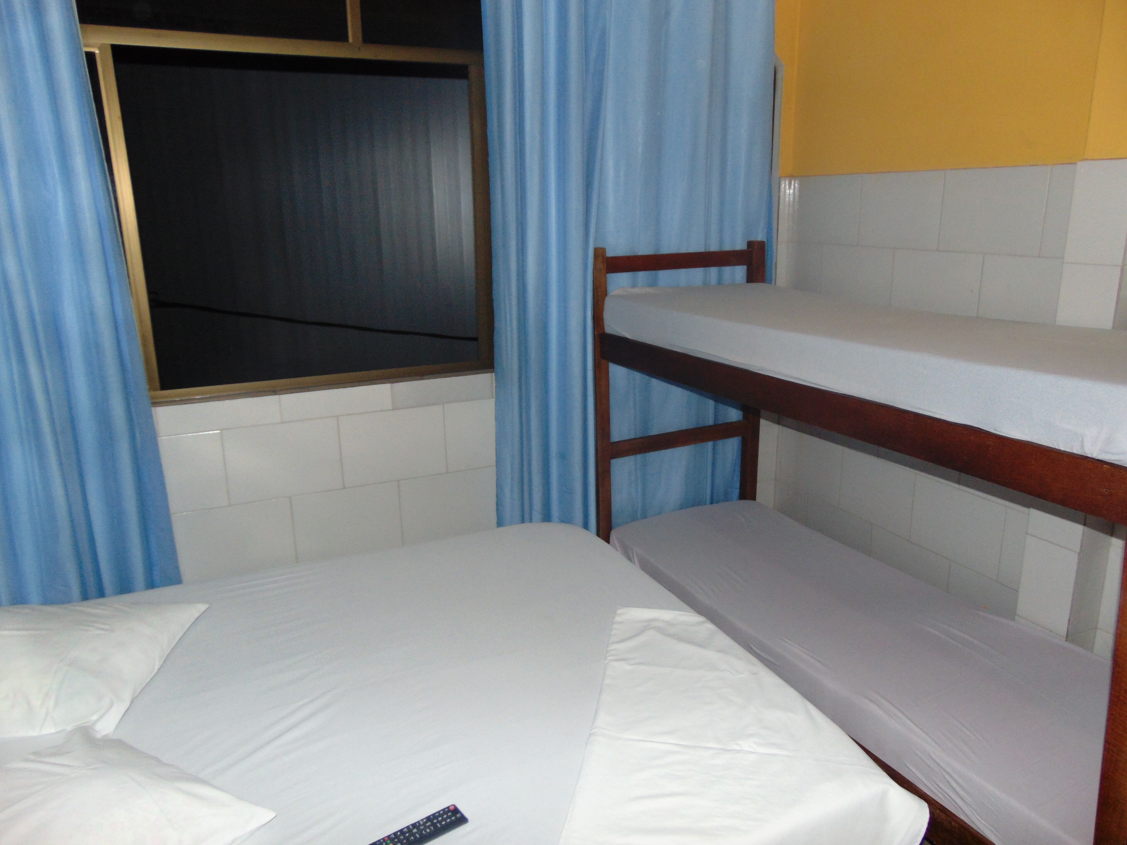 A bunk bed with a wooden frame and white bedding. There is a window with blue curtains, and a yellow wall is visible in the background.