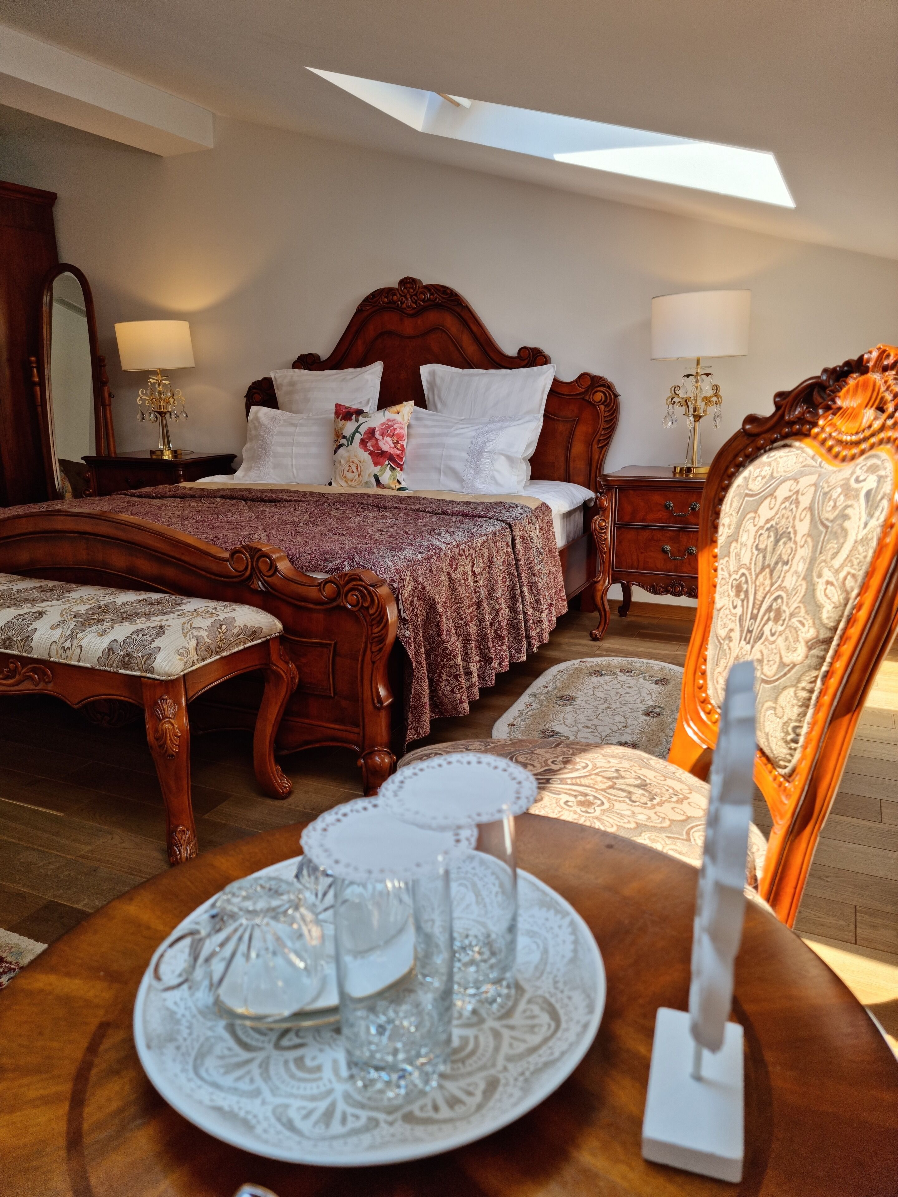 Une chambre à coucher avec un grand lit, une table de chevet en bois, une chaise et une table ornée d’une assiette décorative et de quelques verres.