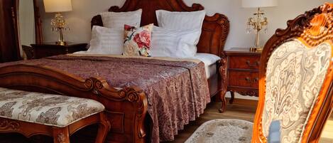A bedroom with a large bed, a wooden nightstand, a chair, and a table with a decorative plate and glassware.