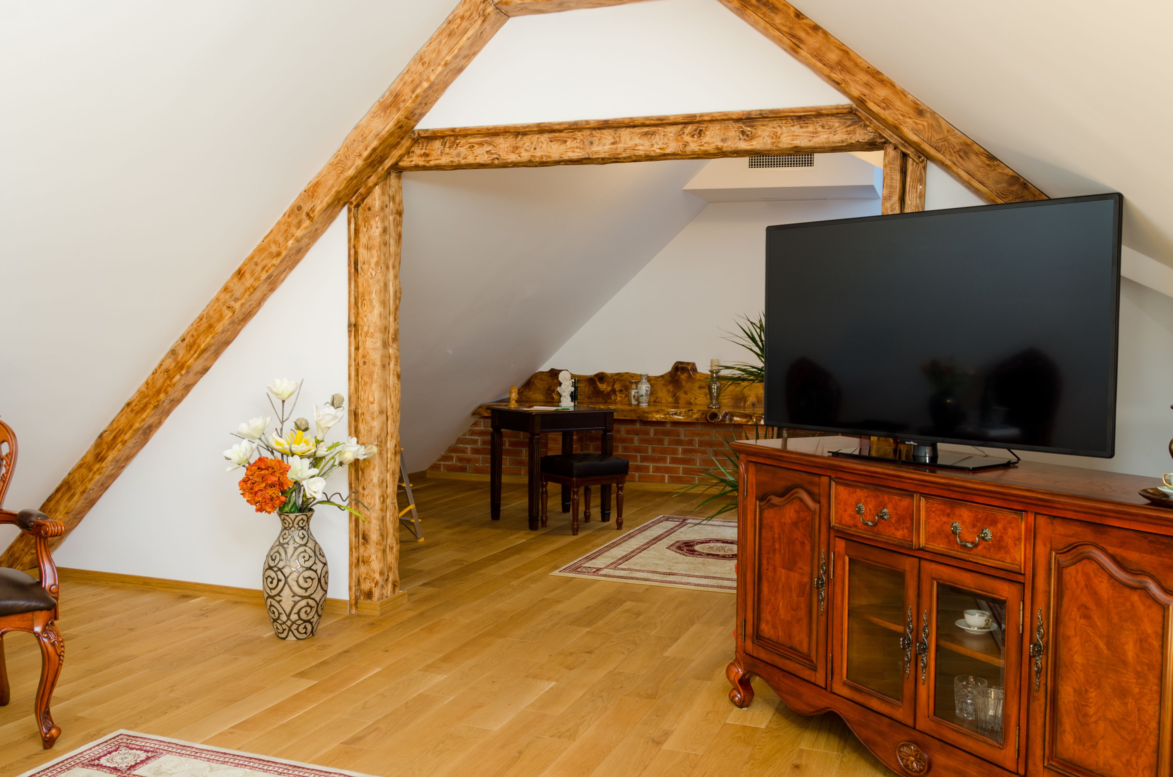 A room with a wooden ceiling, a television on a wooden cabinet, a vase with flowers, and a dining table with chairs.