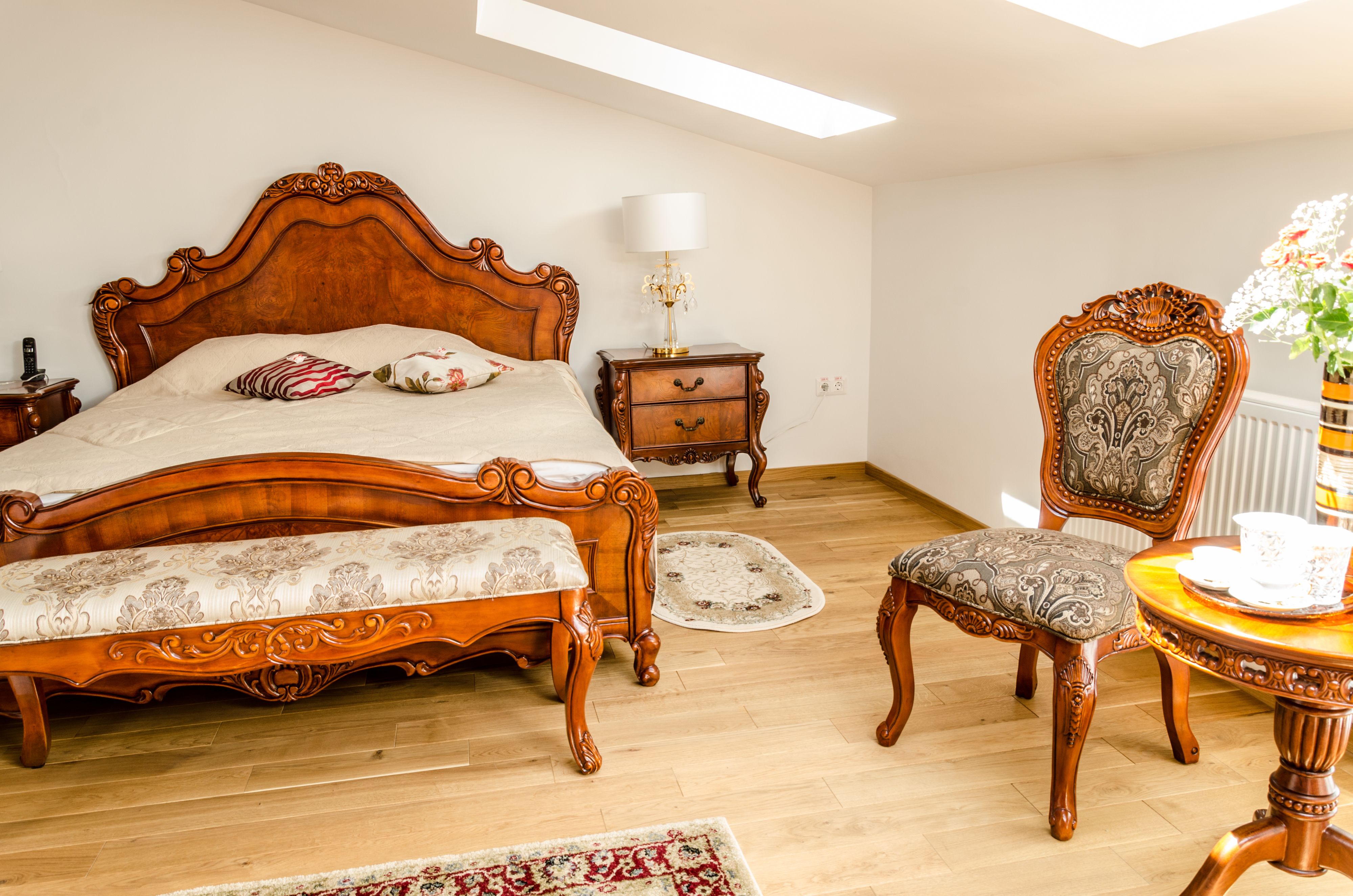 A bedroom with a wooden bed, a nightstand, a chair, a table with a vase, and a window.