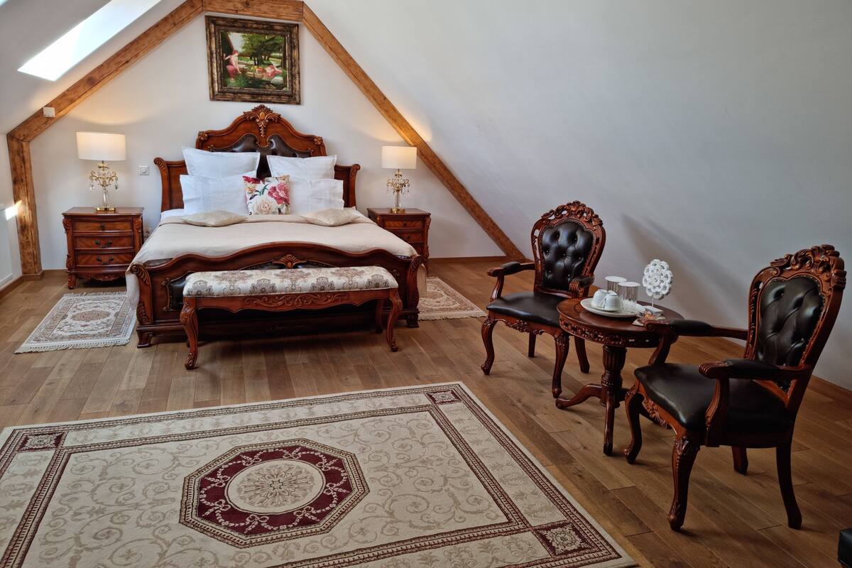 Un dormitorio con techo inclinado, una cama grande, dos mesitas de noche, una silla, una mesita pequeña y un tapete decorativo.