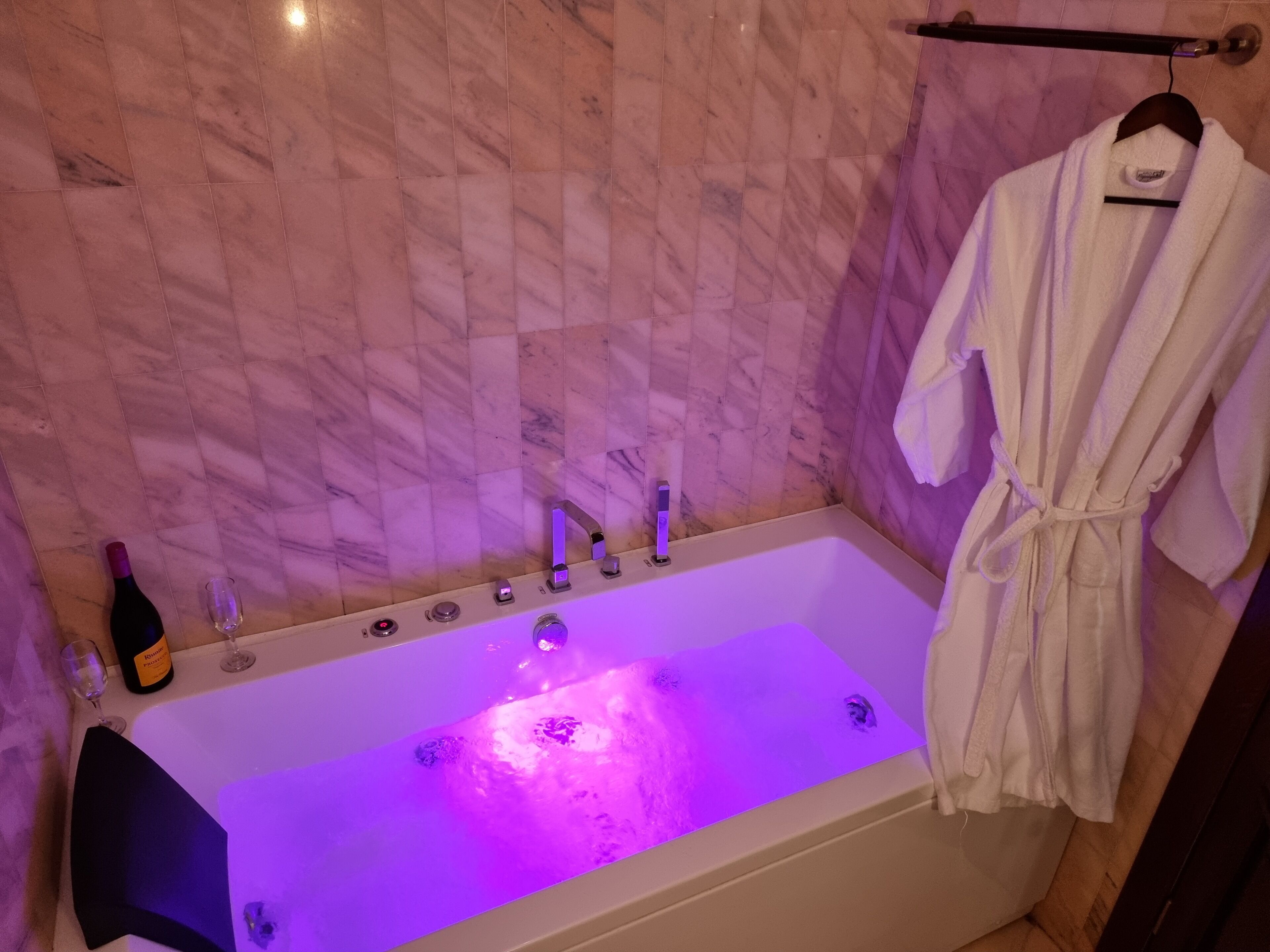 A bathroom with a marble wall, a bathtub with a purple light, a wine bottle, and a robe hanging on a rack.