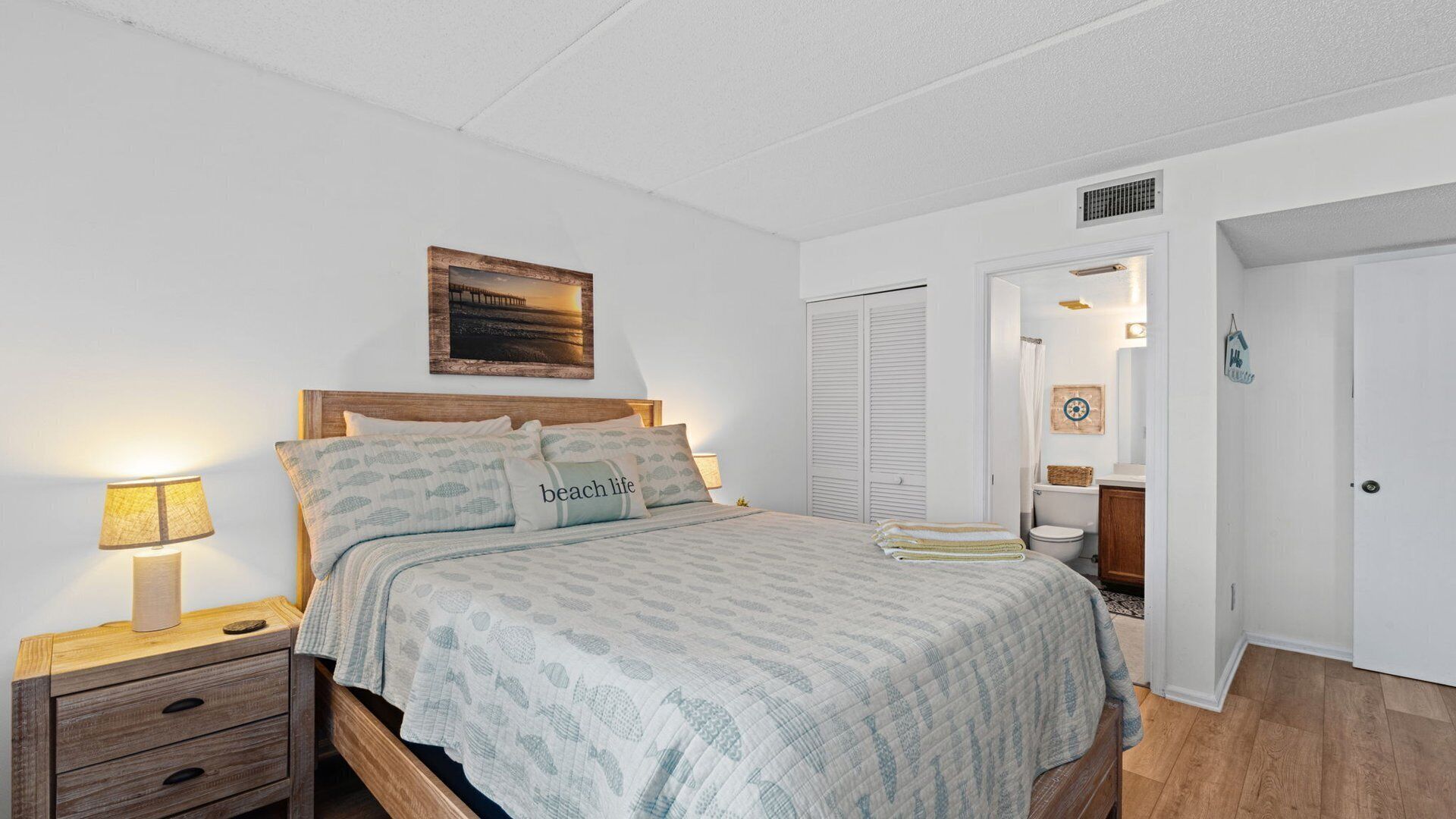 A bedroom with a bed, bedside tables, a nightstand, a framed picture on the wall, and a bathroom door.