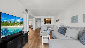 A living room with a flat-screen TV, a sofa, a coffee table, and a dining area with a kitchen in the background. - Poolside St. Augustine Condo – Walk to Beach, Sleeps 4 (St. Augustine)