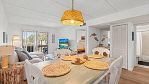 A modern interior with a dining area featuring woven placemats, a white sofa, and a view of the outdoors through large windows. - Poolside St. Augustine Condo – Walk to Beach, Sleeps 4 (St. Augustine)