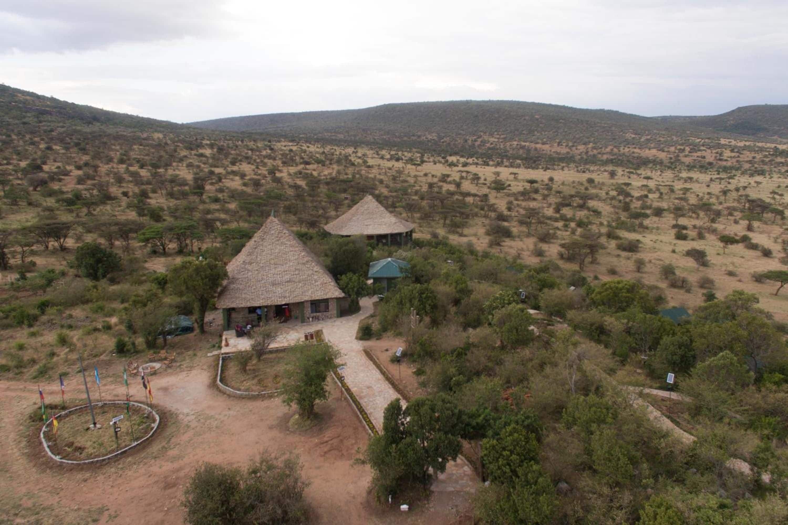 Un entorno rural con dos construcciones de techo de paja, rodeado de vegetación escasa y colinas al fondo.
