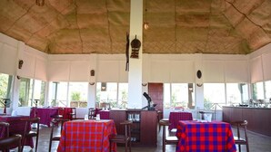 An indoor dining area with tables covered in red and blue checkered tablecloths, wooden chairs, and a high ceiling with a textured design. - La Maison Royale Masai Mara (Maasai Mara)