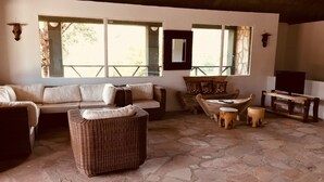 A rustic interior with a stone floor, a sofa, a wicker chair, a wooden table, and a mounted animal head. - La Maison Royale Masai Mara (Maasai Mara)