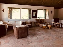 Un interior rústico con piso de piedra, un sofá, una butaca de mimbre, una mesa de madera y una cabeza de animal montada.