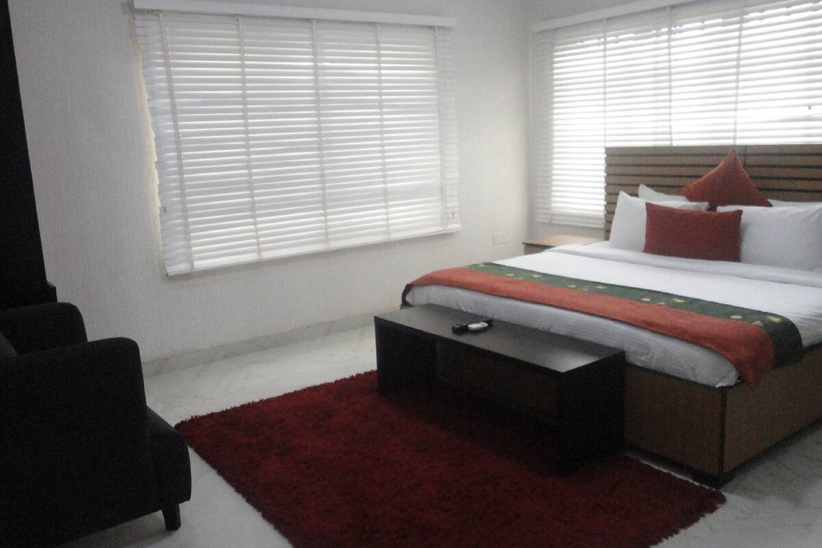 A bedroom with a bed, a chair, a red rug, and white blinds.