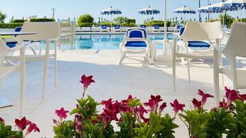 A poolside area with white lounge chairs and blue umbrellas, surrounded by greenery and flowers.
