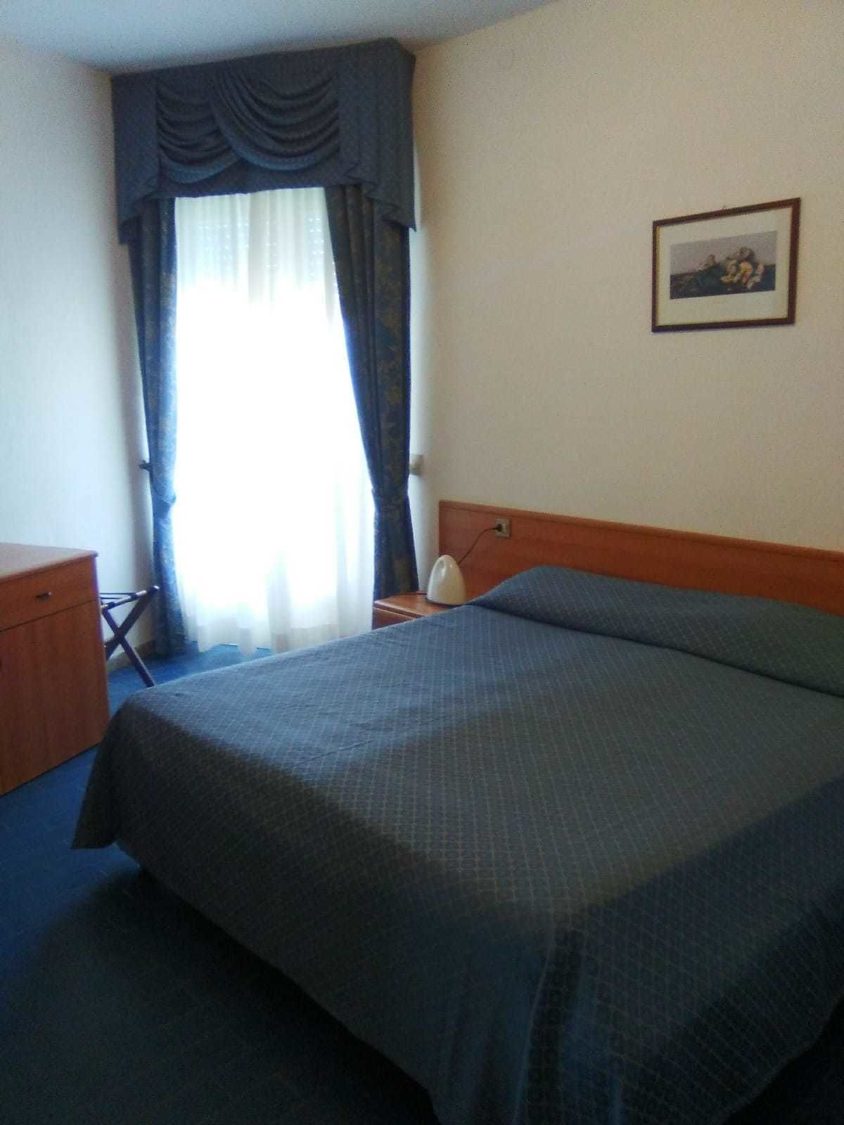 Une chambre d’hôtel avec un lit, un bureau, une chaise, une table de chevet, une lampe et un tableau encadré au mur.