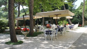 An outdoor dining area with white tables and chairs, covered by beige umbrellas, surrounded by trees and potted plants. - Park Hotel Ravenna (Ravenna)