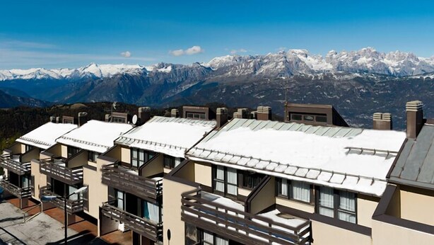 Una hilera de edificios modernos con techos planos, con montañas nevadas al fondo bajo un cielo azul despejado.