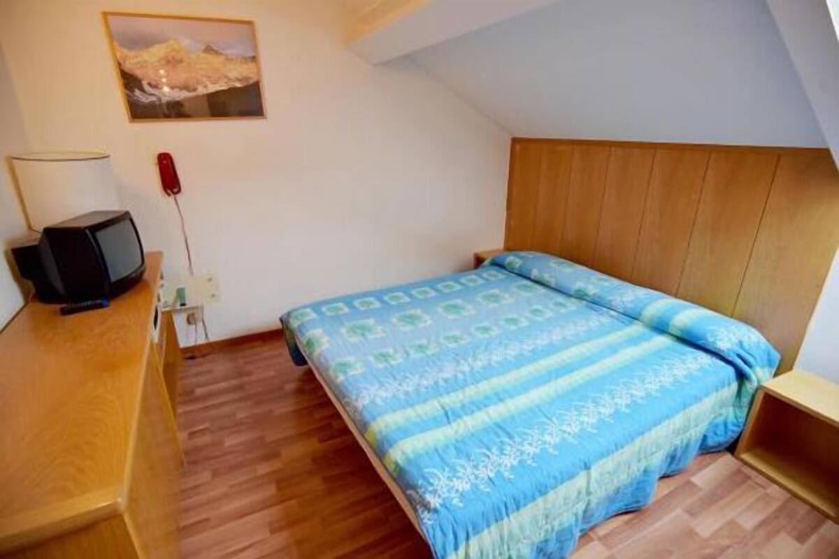 A small, cozy bedroom with a wooden headboard, a single bed with a blue and white patterned bedspread, a wooden desk with a lamp, and a small television.