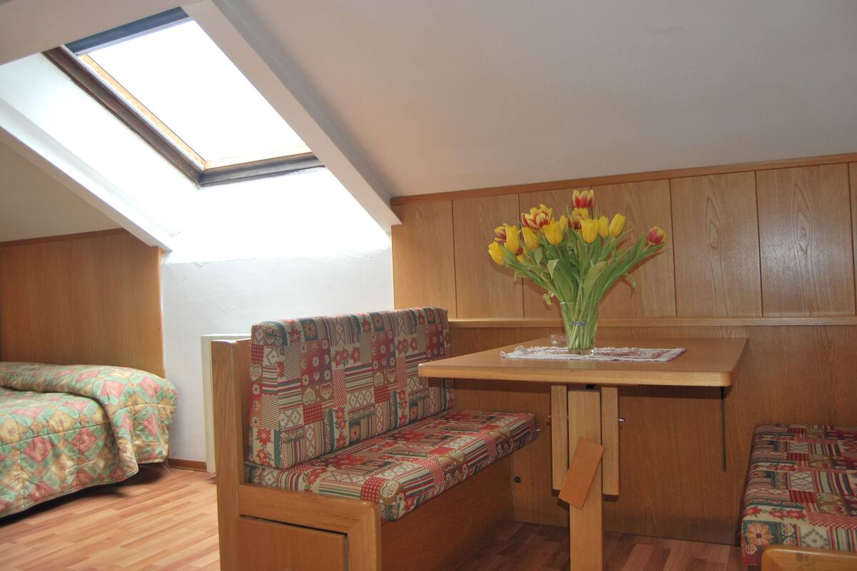 A small, cozy room with a wooden bench and a table, a skylight, and a bed with patterned bedding.