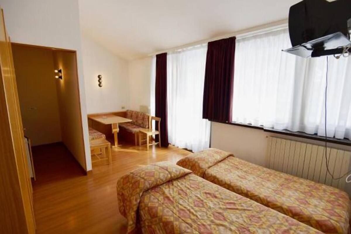 A room with two beds, a small table, and a chair. There is a window with curtains, a radiator, and a door leading to another room.