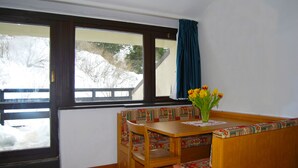 Una habitación con una mesa de madera y un banco, una ventana que ofrece vistas a árboles cubiertos de nieve, y un jarrón con flores amarillas sobre la mesa.