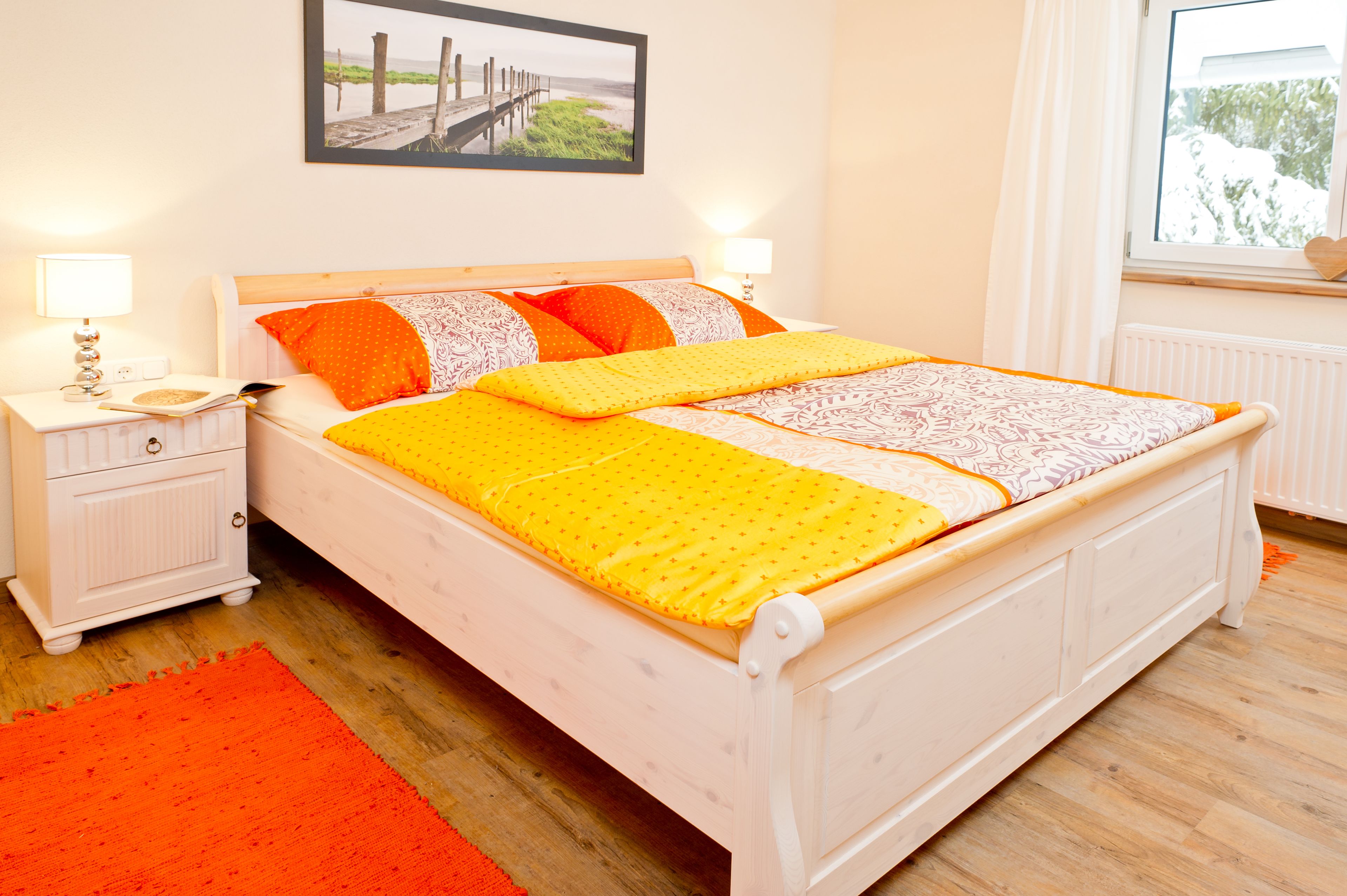 Ein ordentlich bezogenes Bett mit einer gelb-weißen Bettdecke, zwei orangefarbenen Kissen und einem Holzkopfteil. Ein weißer Nachttisch mit einer Lampe und ein kleiner Tisch stehen neben dem Bett. Ein roter Teppich liegt auf dem Boden, und ein gerahmtes Bild hängt an der Wand über dem Bett.