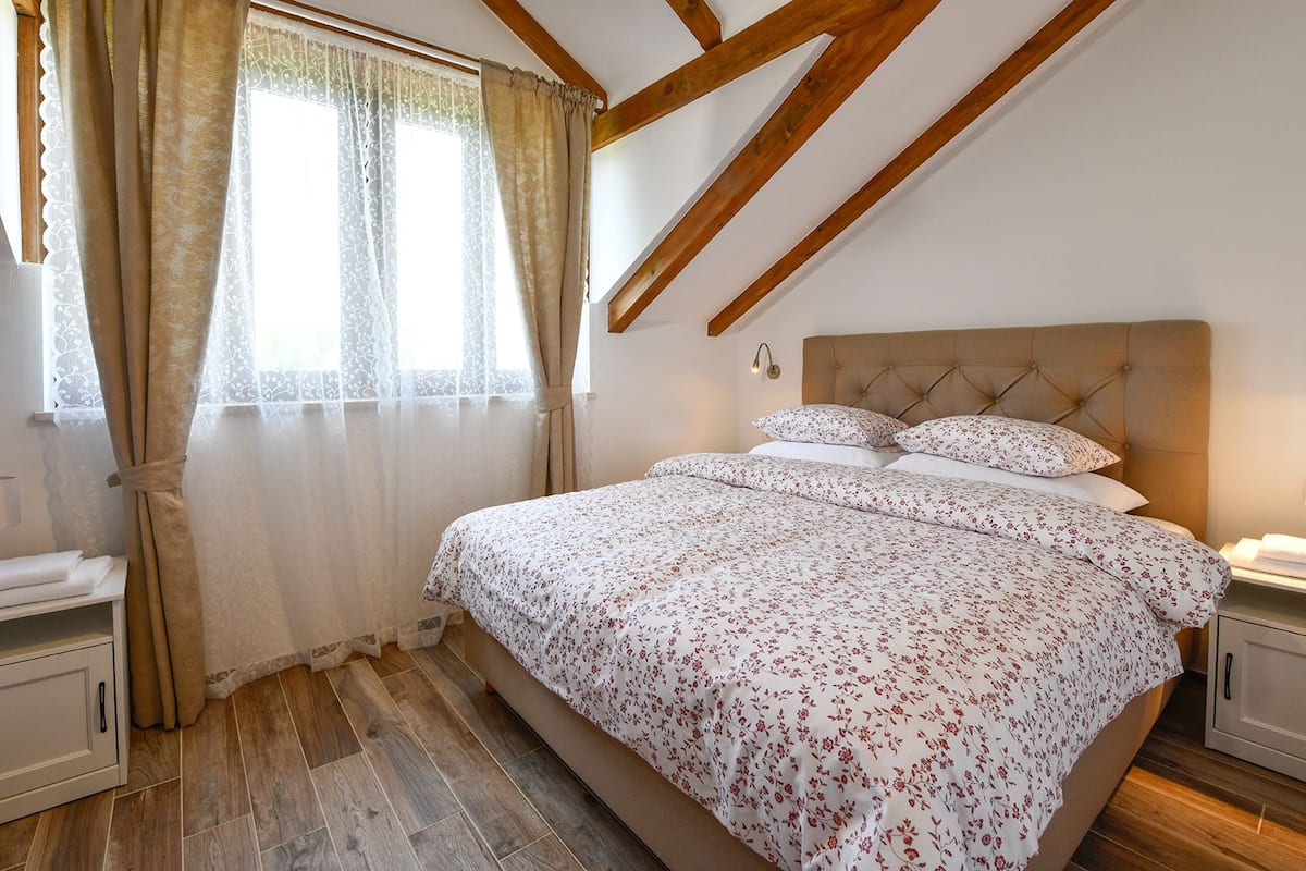 Une chambre avec un grand lit, des poutres apparentes, une fenêtre avec des rideaux et une table de chevet.