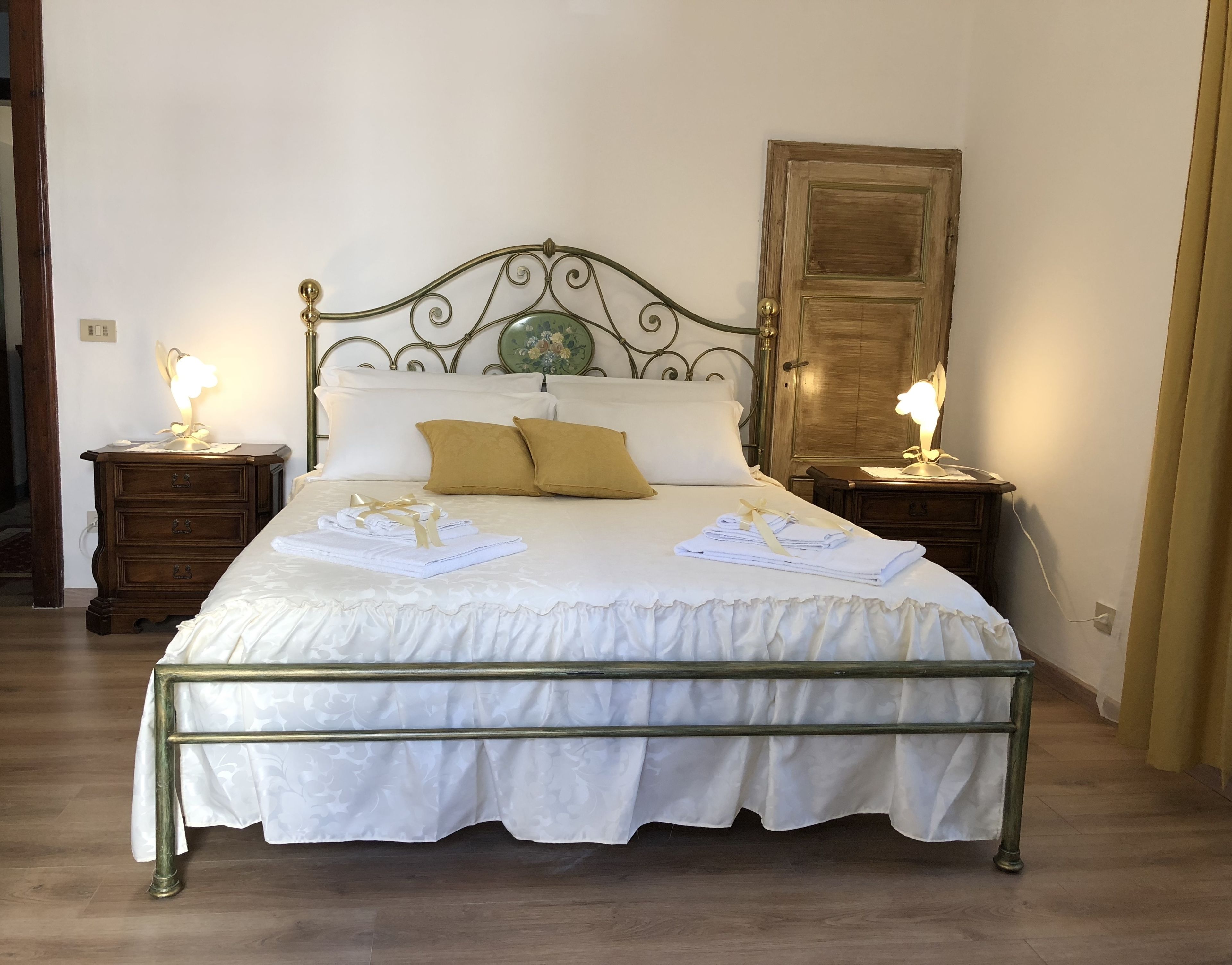 Een netjes opgemaakt bed met witte lakens en twee kussens, twee nachtkastjes met lampen, en een houten deur op de achtergrond.