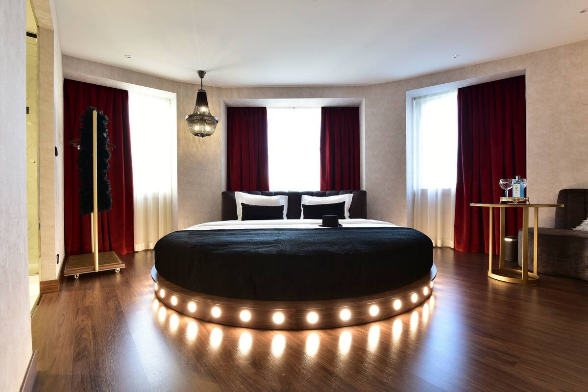 A modern hotel room with a large bed, red curtains, a standing lamp, and a small table with a bottle.