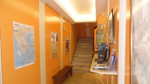 A hallway with orange walls, a wooden bench, a desk with a computer, and a staircase leading to another floor. - Zeus Hostel (Athens)