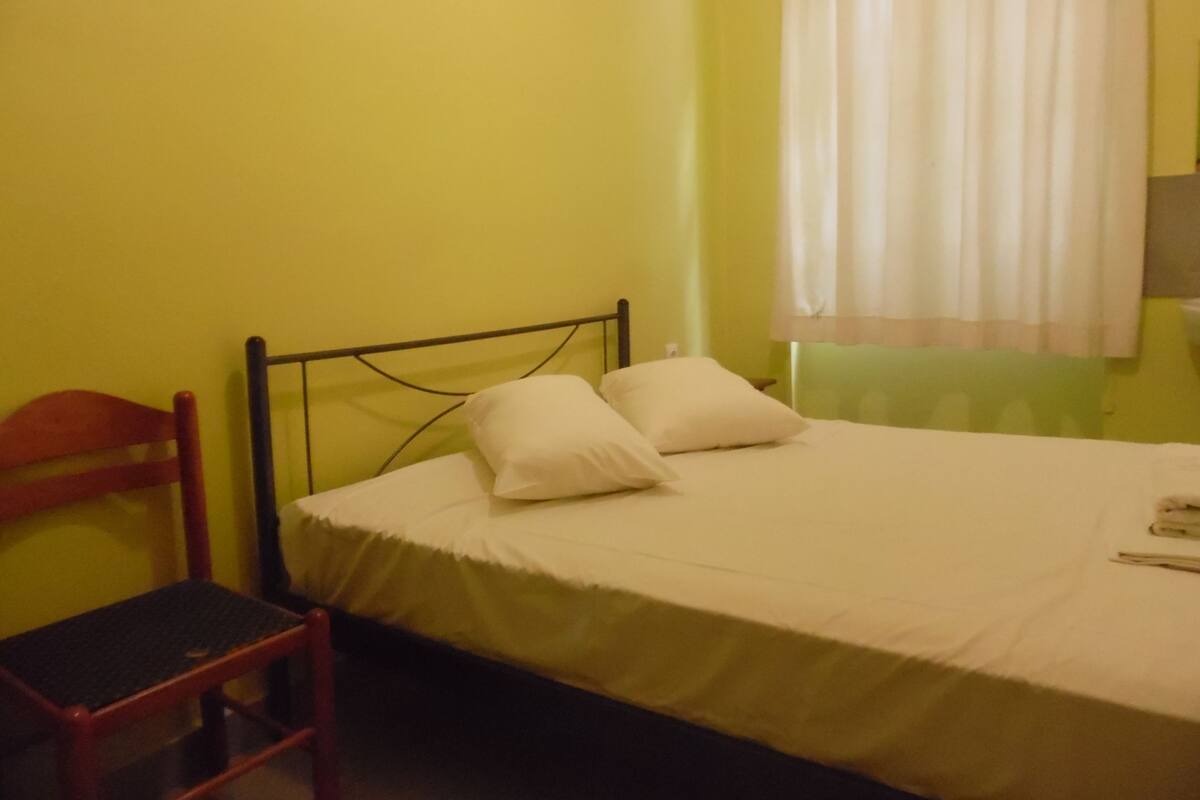 Habitación pequeña con una cama individual, una silla de madera y una ventana con cortinas.