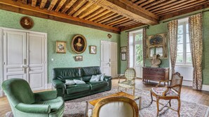 Une pièce avec un canapé vert, une chaise en bois, une table en bois et un tapis à motifs. Il y a deux portes et une fenêtre avec des rideaux.