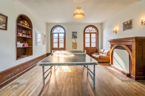 Ein Raum mit einem Tischtennis-Tisch, Holzböden, Rundbogenfenstern, einem Kamin und einem Bücherregal mit Flaschen.