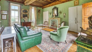 Un salon spacieux avec un canapé vert, un fauteuil vert et une table basse en bois. On y trouve une cheminée surmontée d’un miroir, ainsi qu’un coin repas avec une table et des chaises.