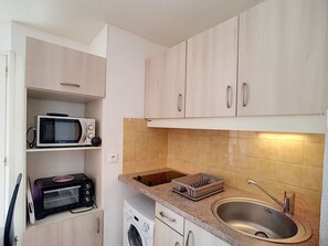 A compact kitchen with a microwave, toaster, and washing machine, a sink with a faucet, and light-colored cabinets. - 1BR Flat with terrace, parking and wifi in Lamalou (Lamalou-les-Bains)
