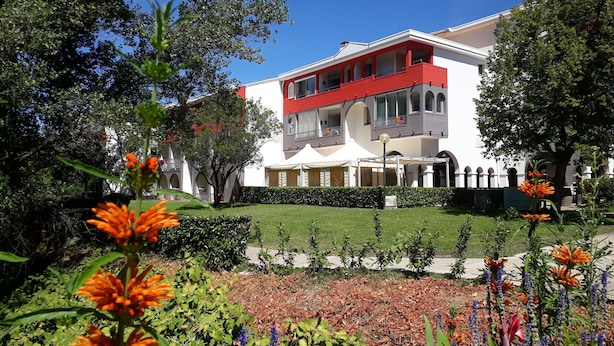 Un edificio de dos pisos con un balcón rojo, rodeado de un jardín cuidado con diversas plantas y flores.
